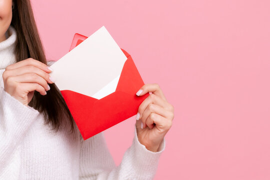 Anonymous woman holding red envelope with blank white card for invitation or romantic message isolated over pink background with copy space