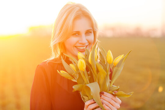 Happy blonde girl laughing and holding yellow flowers bouquet while standing in blurred field during beautiful golden hour