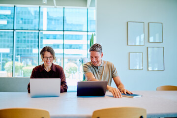 Two cheerful business colleagues working on laptops, sharing a laugh and enjoying successful teamwork in a modern office environment © we.bond.creations