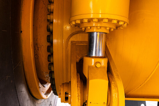 Tight close-up of a yellow-painted industrial hydraulic piston connected to a heavy wheel assembly. Visible components include a chrome piston rod, yellow