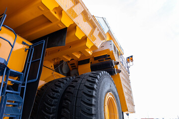 Low-angle view of a massive yellow mining dump truck showing giant treaded tires, heavy metal chassis, ladder access and structural beams. The scene features © NastyaPhoto