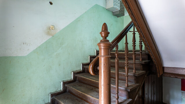 Interior of an old tenement house staircase featuring worn wooden stairs, a brown railing with turned balusters, and a wall painted with a green dado panel under natural daylight.