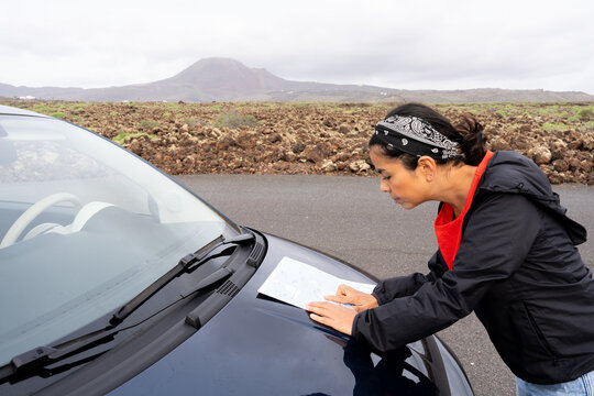 Woman studying a paper map on a car's hood, planning a route through an arid volcanic landscape on a cloudy day