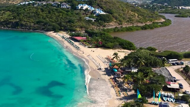 Aerial view of Friar's Bay and Etang Guichard salt pond on the French side of Saint Martin island in the Caribbean. White sand beach organized with beach umbrellas, sunbeds, and people relaxing.
