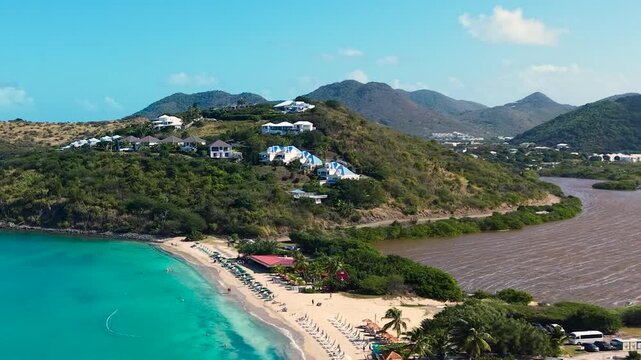 Aerial view of Friar's Bay and Etang Guichard salt pond on the French side of Saint Martin island in the Caribbean. White sand beach organized with beach umbrellas, sunbeds, and people relaxing.