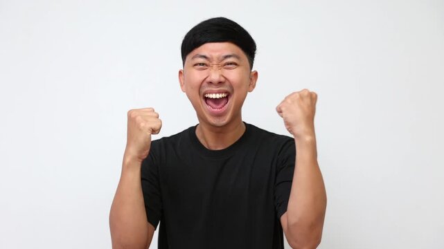 A young Asian man in a black t-shirt celebrating excitedly with raised fists against a white background