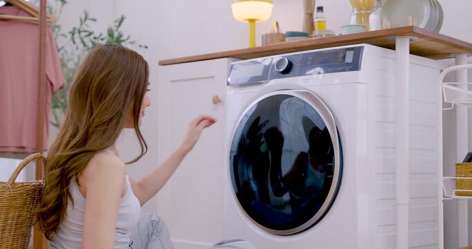 Young Asian woman faces open washing machine, places orange garment into washer drum, raises hand to close door, continuing tidy laundry routine in bright apartment during weekend chore today