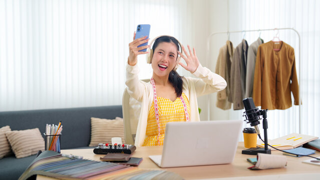 Young Asian female podcaster waving and recording selfie video with smartphone while streaming live from bright home studio workspace with microphone and laptop