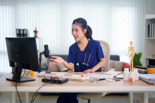 Young Asian female doctor in blue scrubs recording medical podcast with microphone while gesturing at computer monitor in bright modern home office workspace