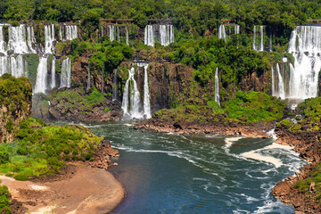 Incredible vista of multiple cascades at Iguazu Falls surrounded by vibrant tropical greenery and a rocky riverbank, perfect for travel, adventure, and nature themes © Yuliko