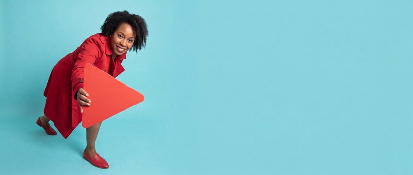 Black woman holding play button sign wearing red coat leaning forward on blue background, happy African American female lifestyle concept banner with copy space for media advertisement marketing