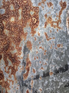 close-up view of a heavily rusted metal surface, likely galvanized iron or steel. Large patches of textured, reddish-brown corrosion contrast sharply against the remaining oxidized silver and grey met