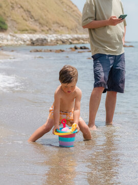 Father is using smartphone while son playing near him at beach 