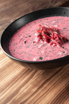 Cold pink beet soup garnished with shredded beetroot and fresh dill, served in a black bowl on a wooden background.