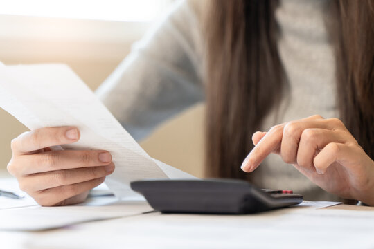Close-up of businesswoman hands holding invoice bill and using calculator to calculate financial report at desk.