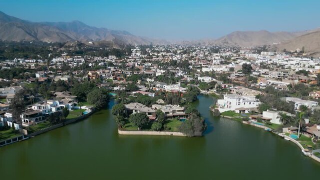 Wide aerial view looking east along the water's edge of the La Planicie private lagoon in La Molina, revealing a row of high end modern white villas, lush landscaping, and distant mountain scenery.