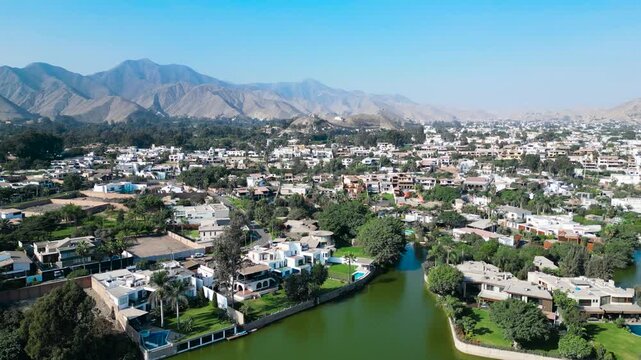 High angle drone establishing shot over the exclusive gated community of La Planicie, highlighting the stark contrast between the lush green properties and the dry desert mountains of La Molina.