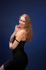Fototapeta premium A woman with a beautiful figure in an elegant black dress poses against a dark blue background.