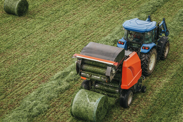Blue tractor with orange baler collecting hay bales in a green field, aerial view showcasing the agricultural landscape and machinery in operation © Emvats
