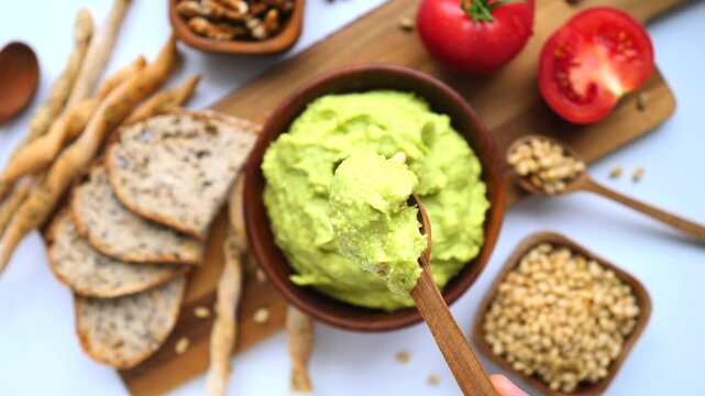 Healthy vegan breakfast table with guacamole in a wooden bowl being stirred by a spoon alongside wholegrain bread, breadsticks, pine nuts, walnuts, and fresh tomatoes on a white background