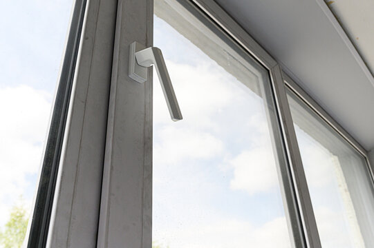 close-up view of a white plastic window handle mounted on a grey PVC frame; the tilted sash reveals a bright sky with clouds through the glass pane