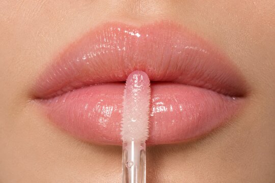 Woman applying pink lip gloss with applicator. Macro shot of lips with shiny cosmetic product. Beauty routine and makeup concept for skincare advertisement and healthy skin care.