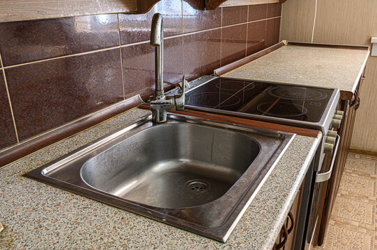 stainless steel kitchen sink with high faucet is installed in speckled granite countertop. adjacent is black glass electric stove, set against brown patterned tile backsplash