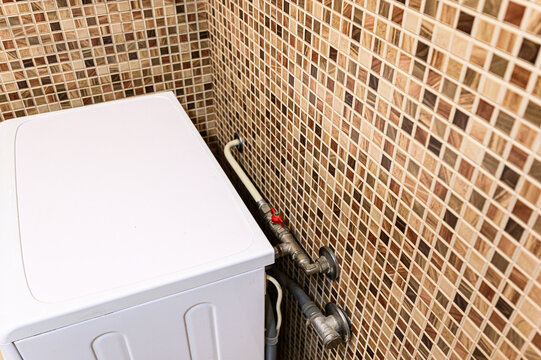 white top-loading washing machine is installed in corner against brown mosaic-tiled wall. visible plumbing includes metal pipes and red shut-off valve