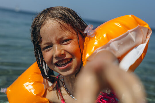Happy little girl with orange arm floaties laughing and reaching toward camera at a Mediterranean beach on a sunny summer day in Sicily, Italy