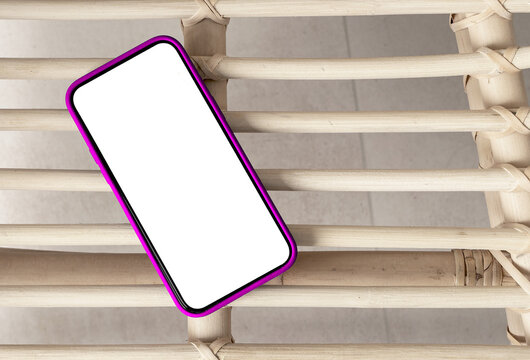 Smartphone with blank white screen and pink case lying on a rattan chair, top view. Mockup template for app or UI design presentation, natural boho interior setting..