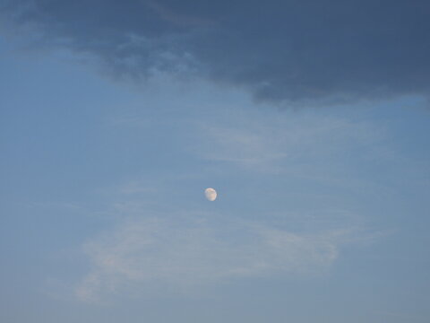 The moon in a blue, daytime, cloudy sky, with a dark cloud looming. Viewed over Kent County, Delaware. 