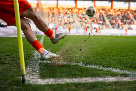 A close-up of a soccer player's legs kicking a ball during a corner kick with turf and dust flying up at a stadium.