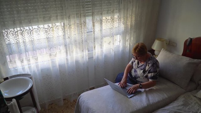 Senior woman using a laptop sitting on the bed at home.