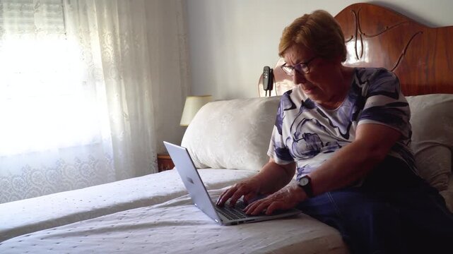  Senior woman using a laptop sitting on the bed at home.