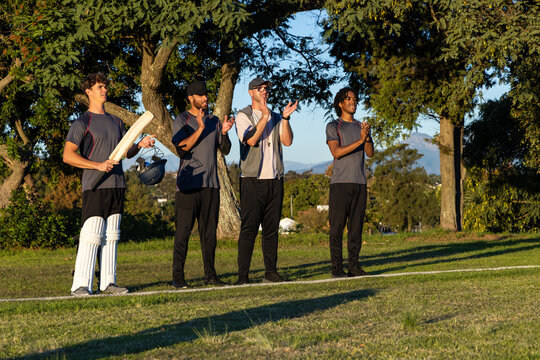 Male cricket teammates in sports shirts standing on field clapping while batsman holding bat, pads