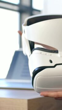 Vertical video: Hand reaching in, person in blue top adjusting white VR headset at desk, inspecting