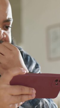 Vertical video: Reacting to phone screen, bearded man in striped shirt holding maroon phone at home