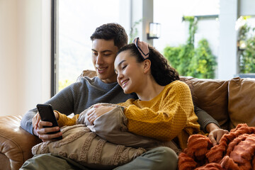 Obraz premium Diverse couple sitting together in living room, leaning and looking at smartphone on leather sofa