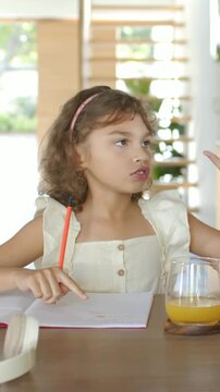 Vertical video: Reading girl in dress tracing open notebook at table, learning with orange pencil