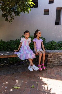Two female children sitting on wooden bench at garden, wearing white sneakers and pink sandals