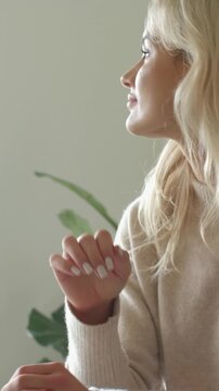 Vertical video: As soft light woman tucking hair tilting right posing in room with beige knit or pla