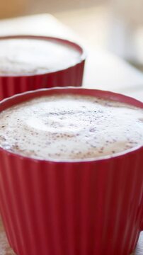 Vertical video: Cam pulling back revealing second red ribbed mug on table by window with cocoa foam