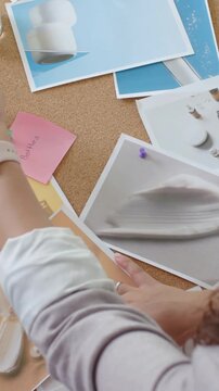 Vertical video: Entering hands smoothing prints on cork board to refine layout with watch and pins