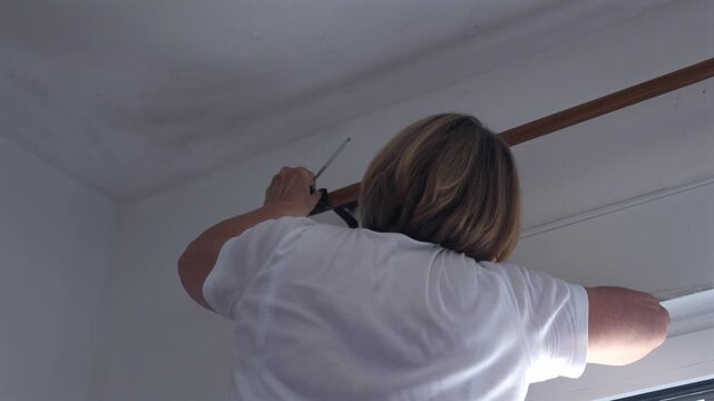 Senior woman using a screwdriver to remove a curtain rod bracket at home. DIY repair, home maintenance and interior renovation concept.