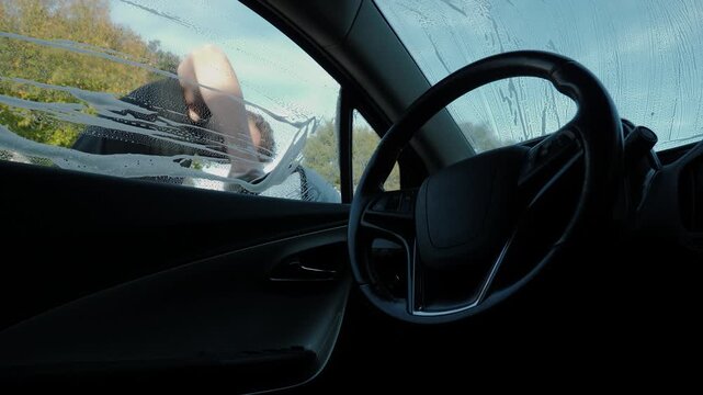Shot from inside a parked car showing the driver-side interior in the foreground and attendant outside wiping soapy water and streaks from the exterior glass. Man wipes exterior glass