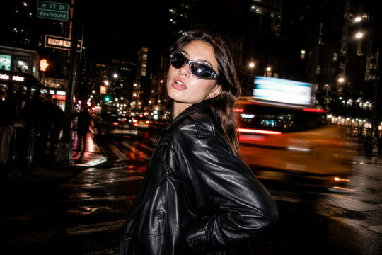 Trendy young woman in leather trench coat and sunglasses on a busy city street at night.