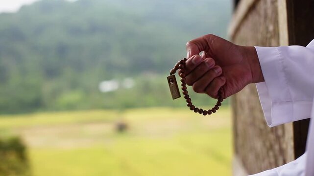 Muslim Indonesian rural man sitting outside his house holding prayer beads tasbih with green rice fields background, traditional spirituality.