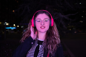 Fototapeta premium A young woman listening to music on headphones by the neon lights at night