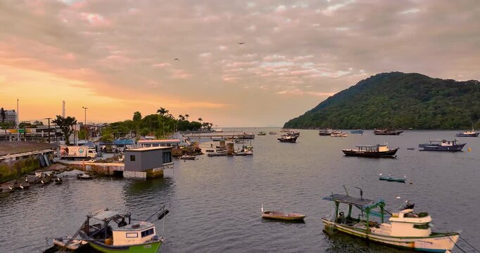 4K drone view of Bertioga fishing harbor at sunset, featuring boats, waterfront, calm waters and coastal landscape on Brazil's coast.