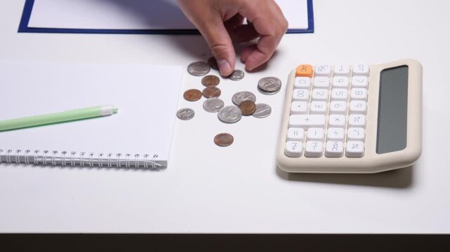 Hand counting scattered coins on a white desk next to a calculator and blank notebook, symbolizing personal finance management, saving money, and budgeting
